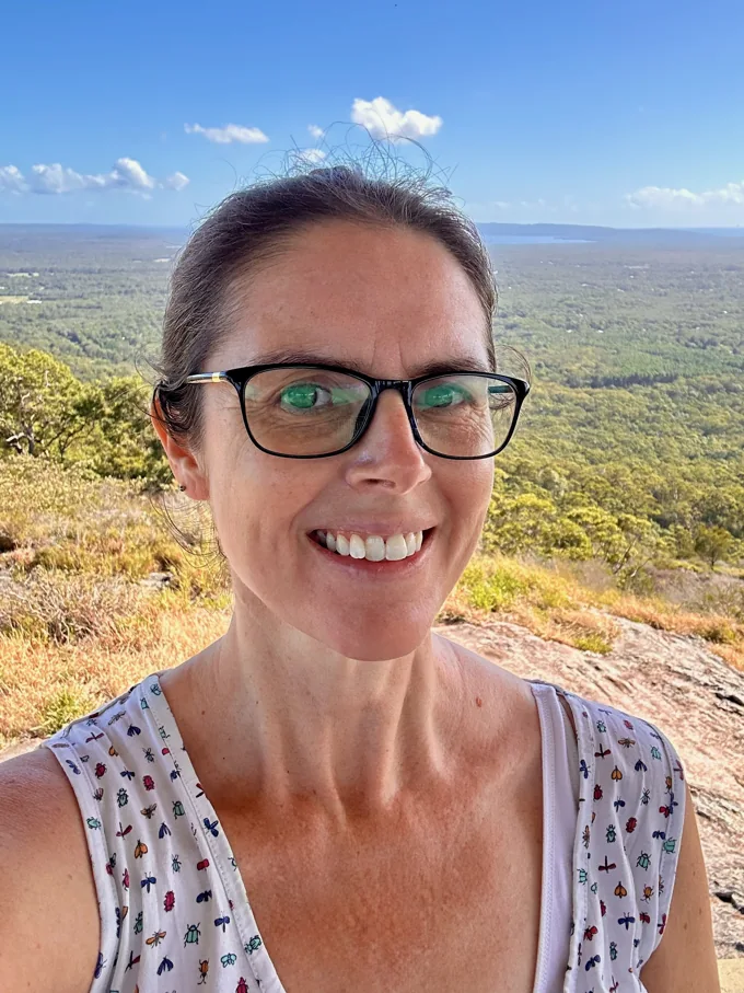 Sally smiling at a lookout, with bushland, coastline and blue sky behind her