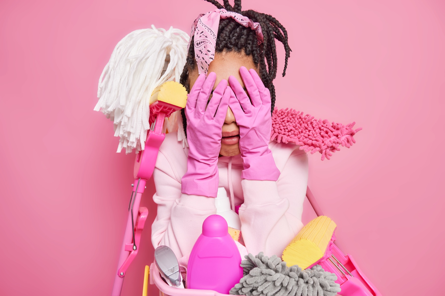 A woman surrounded by cleaning supplies, expressing overwhelm against a pink background